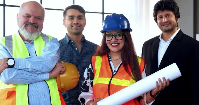 Indian Asian group of civil engineers and architects wearing fluorescent safety jackets, hard hats, holding rolled blueprints, standing confidently with folded arms, smiling, looking at camera