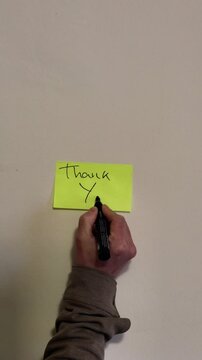 A person writes a thank you note on a piece of paper on a white wall.