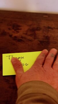 A yellow sticky note with the word "Thank you" handwritten in simple handwriting, stuck on a woden table