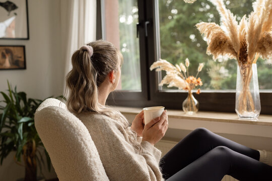 Junge Frau schaut aus dem Fenster, trinkt Tee und legt F&uuml;&szlig;e zum Aufw&auml;rmen auf die Heizung