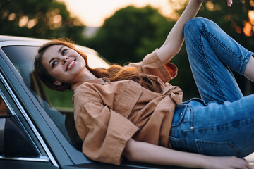 Happy young woman in casual outfit joyfully relaxing on a car hood, featuring a natural outdoor setting during sunset, perfect for lifestyle photography.