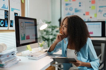 Young businesswoman analyzing financial data on computer screen and holding clipboard with pen in...
