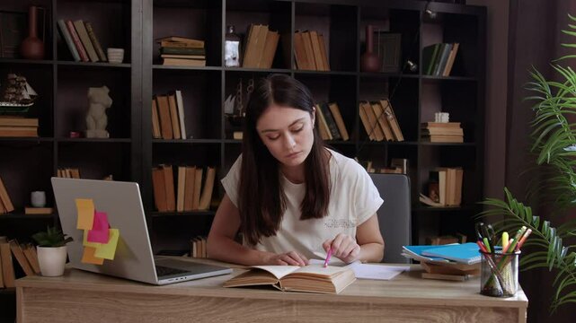 Female student sitting in library doing homework and preparing for college exam test. Young woman writing notes in notebook from textbook carefully reading information from book writing on paper