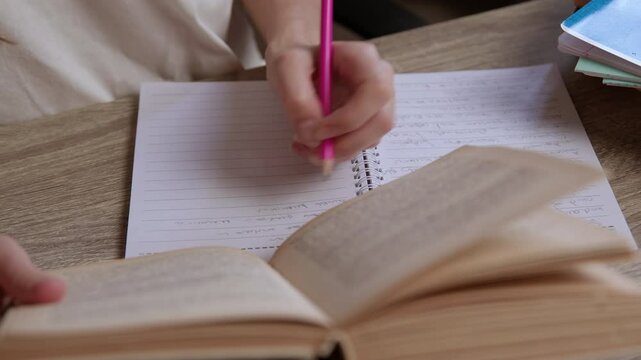 Close-up image of a female student studying and summarizing information from a book and taking notes on paper. The left-handed girl is reviewing several ideas from the book and writing in a notebook