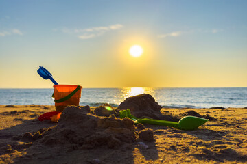 Childrens beach sand toys bucket and shovel on the sea coast on background calm blue ocean water in the sunset rays. Summer vacation, kids fun activities, building sandcastles play tools.