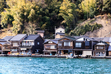 Fototapeta premium Traditional Wooden Boat Houses Along the Waterfront in Ine, Japan, with Tree-Covered Hills in Soft Natural Light