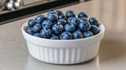 A white bowl filled with fresh blueberries sits on a countertop, showcasing their vibrant blue color and healthy appeal.