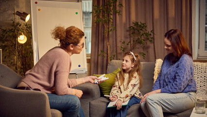Psychologist offering a pencil for a little girl, child sitting on couch next to her mother....