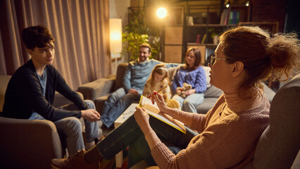 Psychologist takes notes, listening to teen boy expressing his thoughts and feelings, family members sitting in background. Concept of psychology, assistance, family therapy session