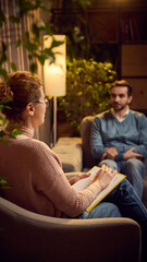 Female psychologist writing notes during therapy session with male client sitting on a couch,...