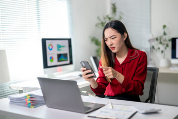 A woman in a red shirt is looking at her cell phone while sitting at a desk