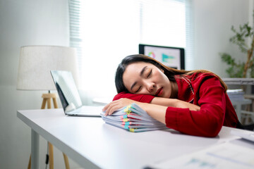 A woman in a red shirt is sleeping on a desk with a laptop and a stack of papers