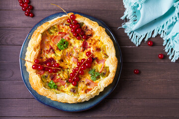 Quiche with chunks of ham, creamy brie and vegetables on blue plate on brown wood background. View from above, top view