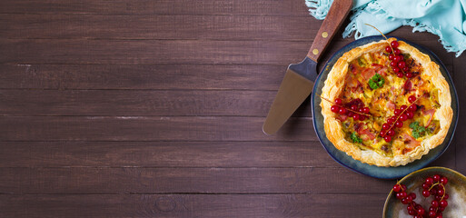 Quiche with chunks of ham, creamy brie and vegetables on blue plate on brown wood background. View...