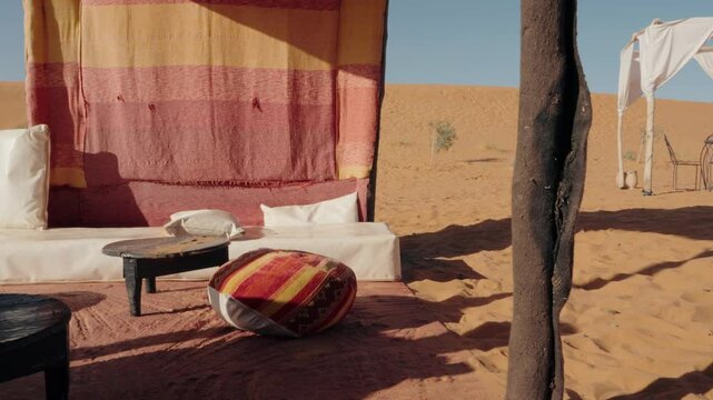 Relaxing desert camp in Erg Chebbi, Sahara Desert, Merzouga, Morocco. A comfortable lounge area under a tent provides shade from the midday sun. Traditional Moroccan furniture and colorful textiles