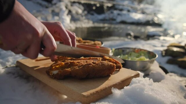 Close up of young man cutting delicious schnitzel on wooden board near beautiful mountains river, snow covered forest and bonfire at winter time. Cooking outdoors concept