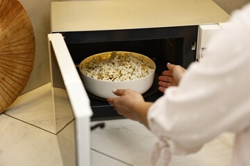 Woman taking bowl with popcorn out of microwave oven at white marble countertop in kitchen, closeup