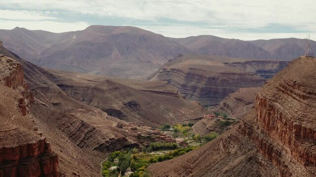 Breathtaking aerial view of a small village nestled in the Dades Gorge, Morocco. The lush greenery contrasts with the rugged, layered rock formations of the High Atlas Mountains,