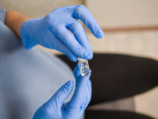 Medical professional wearing blue nitrile gloves handling glass vial with clear injectable liquid, precise technique and careful attention to glass vial in sterile healthcare setting. 