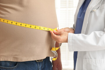 Nutritionist measuring overweight man's waist with tape in clinic, closeup