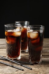 Refreshing cola with ice cubes in glasses and drinking straws on wooden table against black background