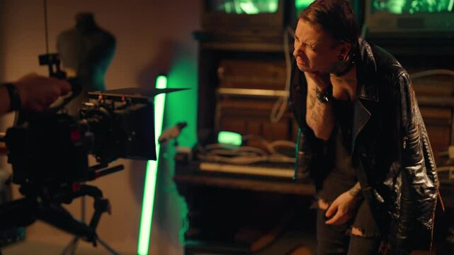 Professional shooting in studio, cameraman and actress playing role of rockstar. Young adult woman with tattoos and piercing on body scratching her neck, autoaggression, self-harm and destruction