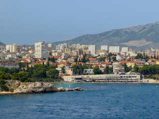 Split, Croatia - July 1, 2024: Southeast of harbor. Kavala rocky point with Bacvice Beach and hotel, restaurants, and clubs long building along boardwalk. Tall buildings cityscape in back in front of 
