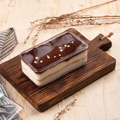 A creamy dessert with chocolate topping sits on a wooden board, complemented by dried herbs on a textured surface