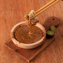 Chopsticks hold a dumpling above a small wooden bowl filled with rich dipping sauce alongside fresh lime halves on a wooden surface