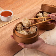 Hands skillfully using chopsticks to pick up dumplings from a bamboo steamer, accompanied by a small dish of dipping sauce