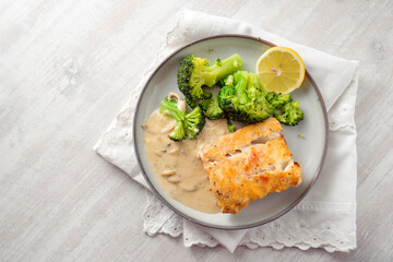 Fried cod fish fillet with broccoli, sauce and a slice of lemon on a gray plate and a white painted wooden table, healthy meal for a low carb diet, high angle view, copy space