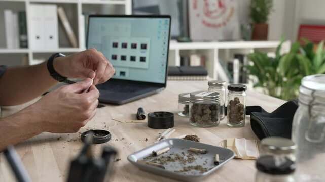 Asian male creative industries worker, rolling cannabis joint in an office, surrounded by a relaxed ambiance with cannabis equipment and accessories on the table.