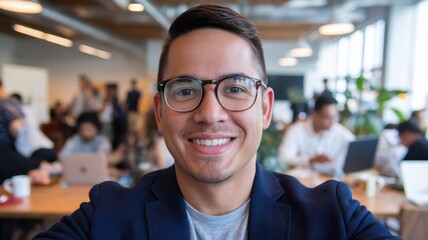 Fototapeta premium A young Hispanic man with glasses smiles warmly at the camera in a bustling coworking space filled with people.
