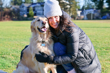 Adult woman wearing beanie cap and gloves for cold weather next to her friendly pet golden retriever dog outside on green grass. 
