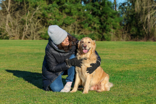 Adult woman wearing beanie cap and gloves for cold weather next to her friendly pet golden retriever dog outside on green grass. 