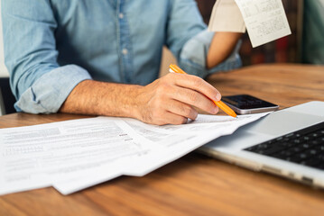 Man in a denim shirt calculating taxes with receipts and forms on a wooden desk. Tax preparation...
