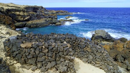 Volcanic rocks are forming a wall in Charco del Palo, Lanzarote, Canary Islands, Spain, near a beautiful blue sea with waves crashing on the rocky shore