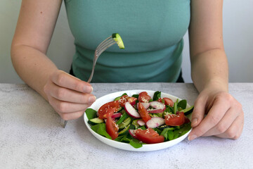 Woman hands cooking fresh healthy organic salad of spinach, cucumber, tomato and radish in white bowl on table top view on grey background.