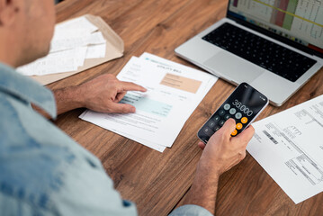 Man calculating utility bills using a smartphone calculator and reviewing documents on a wooden...