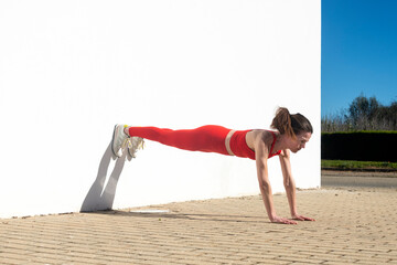 Athletic woman female performing plank exercise outdoors against white wall,