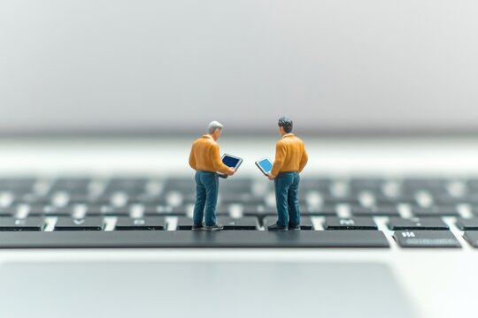 Two miniature figures stand on a laptop keyboard, each holding a tablet, symbolizing modern collaboration.