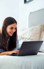 Naklejka premium One laxy and cute young woman chatting online at home laying on bed with a laptop. Morning light in background. Concept of indoor leisure activity with computer or smart working