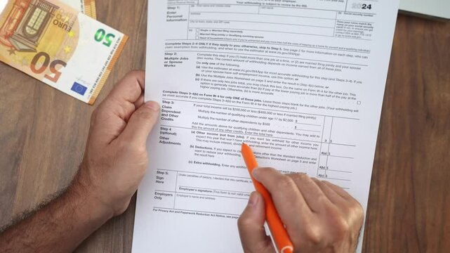 Male accountant analyzes details on W-4 tax form with pen, man works at table with euro bills and laptop ensuring document complies with tax regulations slow motion