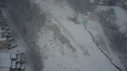 A breathtaking aerial view of a snow-covered neighborhood, featuring rows of houses, parked cars, and winding roads. Bare trees and light tire tracks add charm to the serene winter scene.