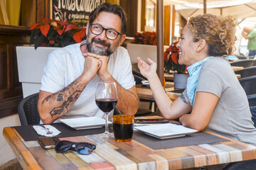 Tourists enjoying their vacation at a cozy outdoor restaurant, sitting at a table, smiling and chatting over wine and cola as they relax and wait for their lunch to be served in the sunny environment