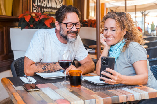 A 48-year-old couple laughs and chats together at an outdoor restaurant table, enjoying a peaceful and relaxed lunch as the woman shares funny content from her phone with her husband under the sun. - Powered by Adobe