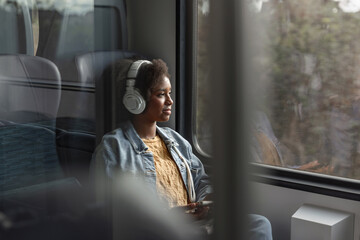Young African American woman comfortably sitting by the train window, watching the beautiful scenery and enjoying music through her headphones. © 24K-Production