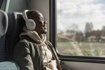 Smiling African American man enjoying relaxing and comfortable train travel, sitting by a window, and listening to music through headphones.