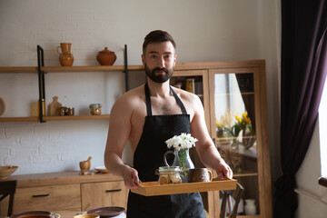 Handsome attractive man standing in kitchen with tray with flowers and coffee. High quality photo.