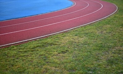 Red running track with blue sports field in left side and green grass in right side, sunny day